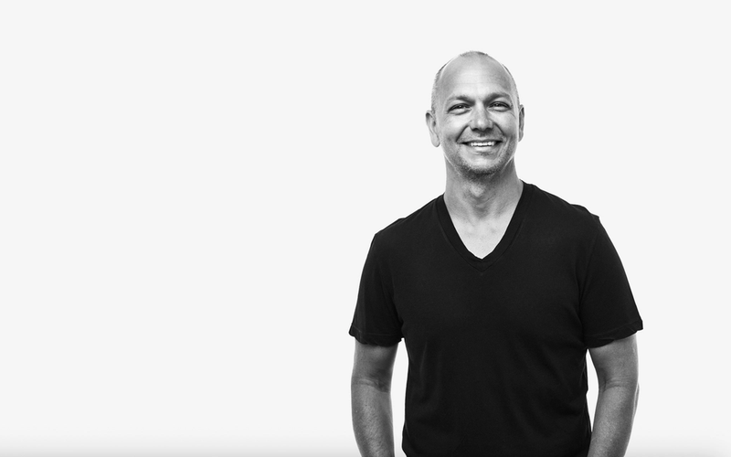 A black-and-white photo of Tony Fadell, smiling, wearing a short-sleeved, V-neck T-shirt. He is standing casually with one hand in his pocket against a plain white background.